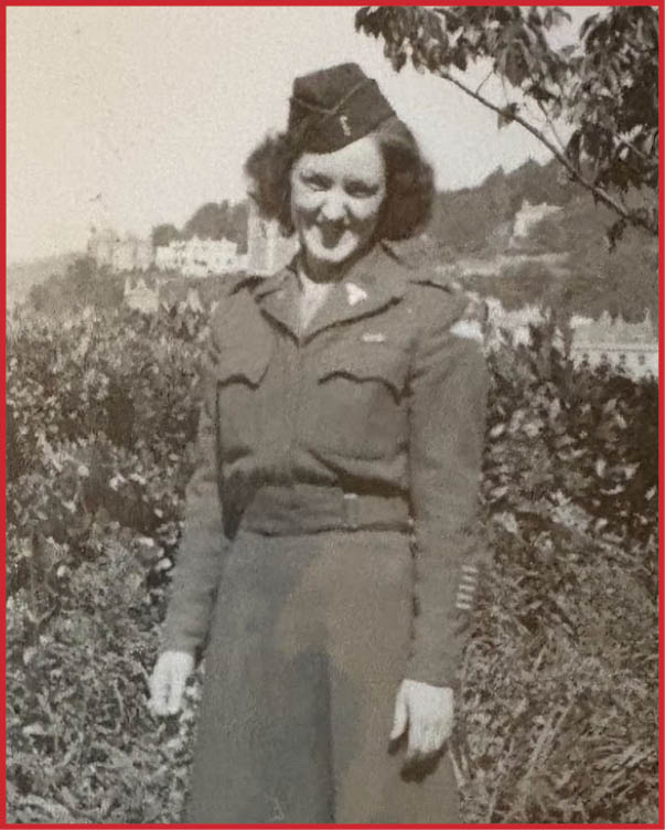 A person wearing a vintage military uniform stands on a path next to dense foliage with a distant view of buildings and trees in the background. Description generated by AI
