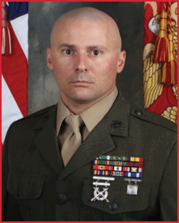 A person in a military uniform with medals and ribbons stands in front of American and Marine Corps flags. Description generated by AI