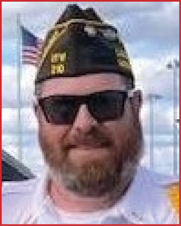 A person wearing sunglasses and a military-style hat with insignia stands outdoors with an American flag and light poles visible in the background. Description generated by AI