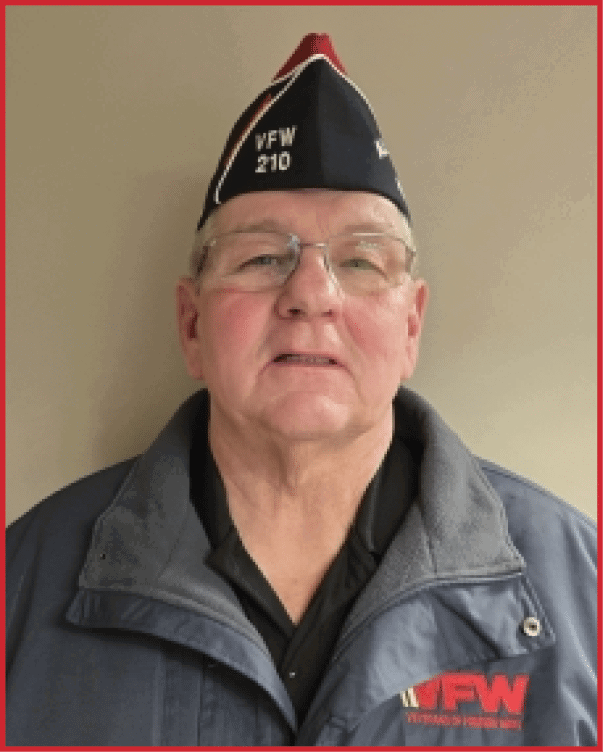 A senior wearing glasses and a dark jacket with a VFW Veterans of Foreign Wars logo stands against a plain background. Description generated by AI