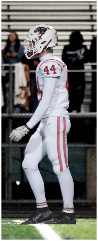 A person wearing a white football uniform with the number 44 stands on a field near the sideline with empty bleachers and a few spectators in the background. Description generated by AI