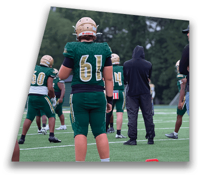 A person wearing a green football jersey with the number 61 and a gold helmet stands outdoors near another person in a similar helmet and a person in a dark hooded sweatshirt. Description generated by AI