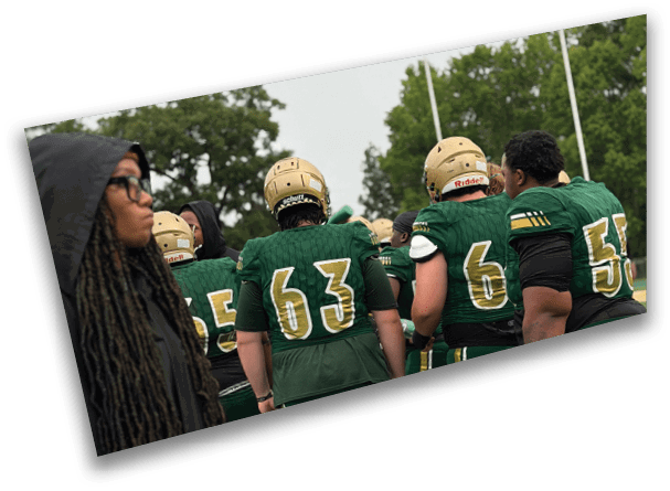 People wearing green sports uniforms and helmets are gathered outdoors near trees under a cloudy sky. Description generated by AI