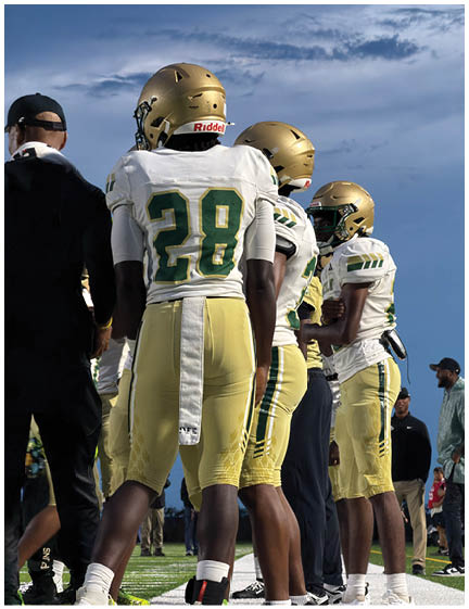 Three people wearing gold helmets and white and gold football uniforms stand on a field under a partly cloudy sky. Description generated by AI