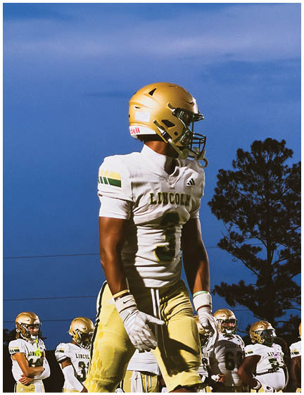 A group of people wearing gold helmets and white sports uniforms are standing on a field under a blue sky, with one person prominently in the foreground looking to the side. Description generated by AI