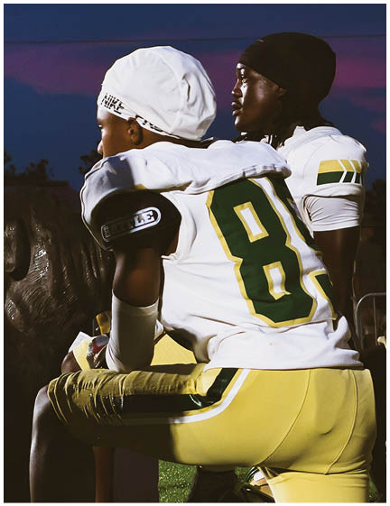 Two people wearing football uniforms with helmets and pads are sitting closely together, one with an arm around the other, against a dark outdoor background. Description generated by AI