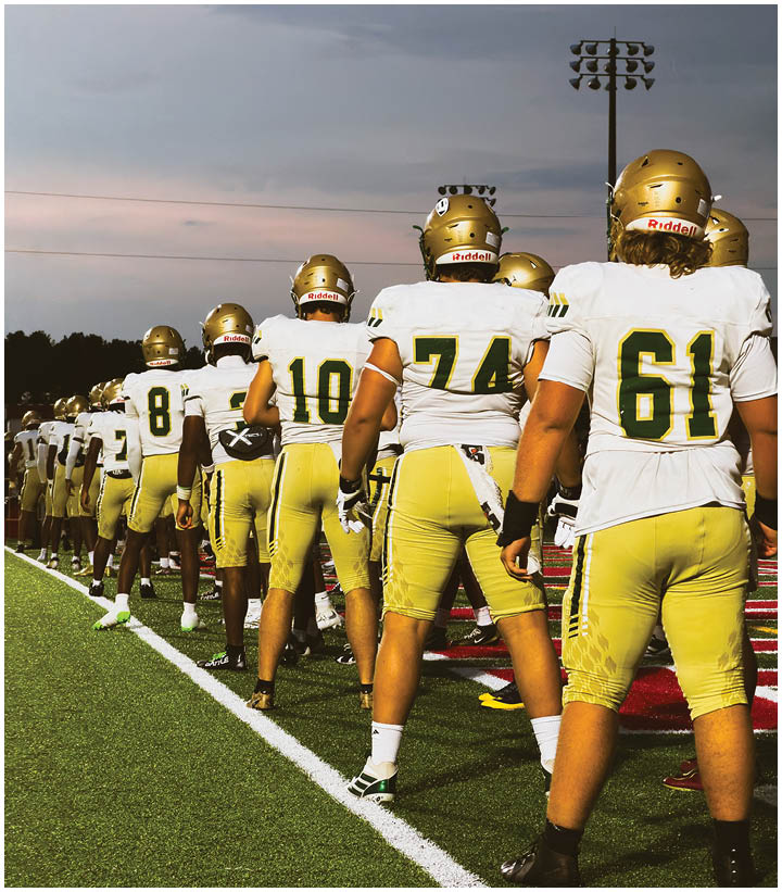 A group of people wearing yellow football pants and white jerseys with gold helmets stand in a line on a football field under a cloudy sky. Description generated by AI