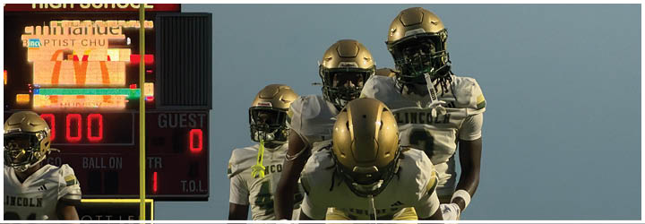 Four football players wearing gold helmets and white uniforms are positioned in front of a scoreboard at Crestview High School during a game. Description generated by AI