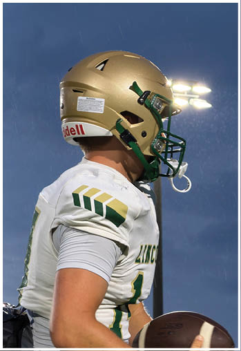 A person wearing a gold football helmet and white jersey with green and gold accents is standing outdoors under stadium lights during dusk or evening. Description generated by AI