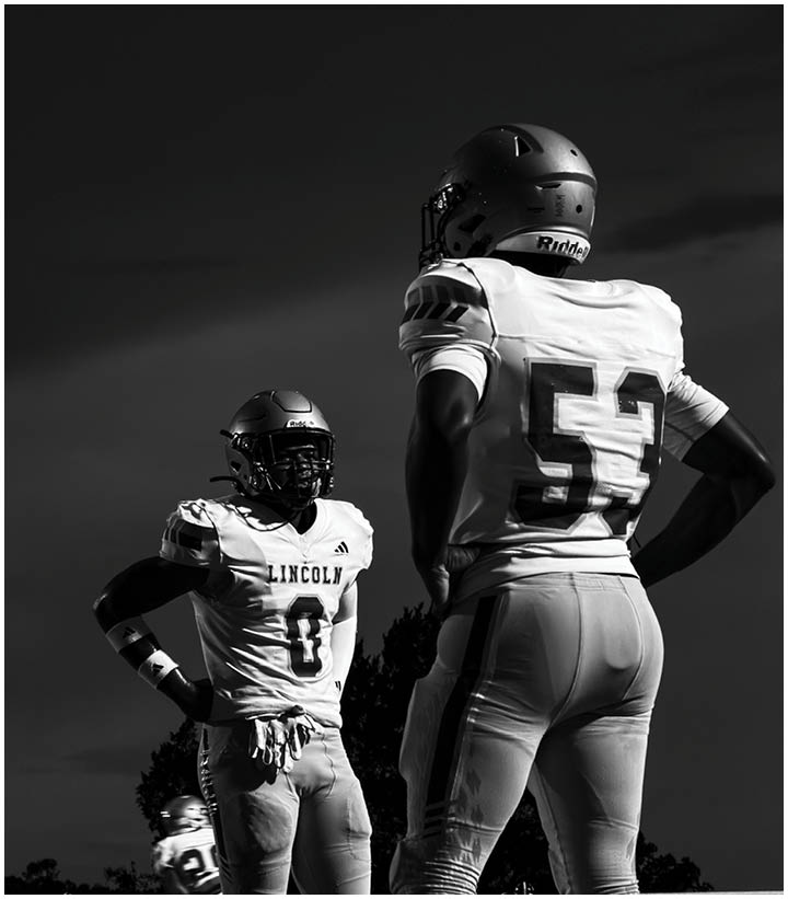 Two people wearing football helmets and uniforms stand facing each other outdoors under a dark sky. Description generated by AI