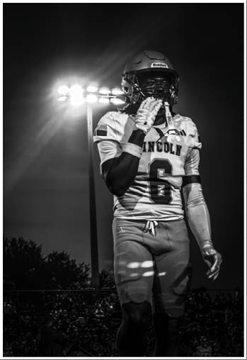 A person wearing a football uniform with the number 6 and the word Lincoln on the jersey stands on a field under bright stadium lights at night. Description generated by AI