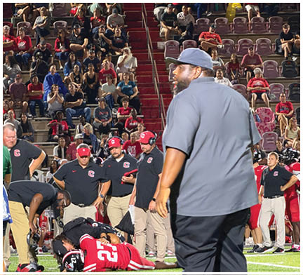 A group of people wearing sports uniforms and casual clothing are gathered in a stadium with bleachers and a building labeled “HOME" in the background during a nighttime event. Description generated by AI