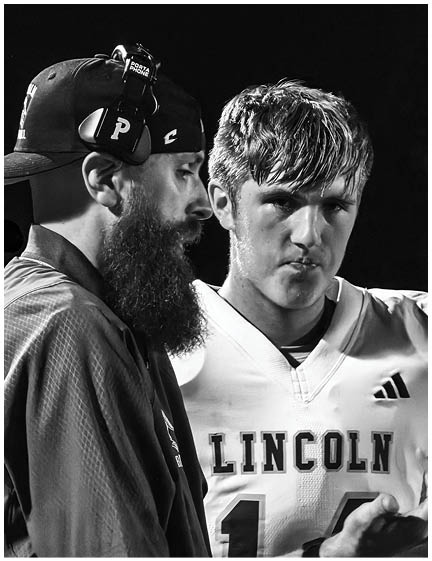 A person wearing a headset and cap is speaking closely to a person in a Lincoln sports jersey who has a serious expression. Description generated by AI