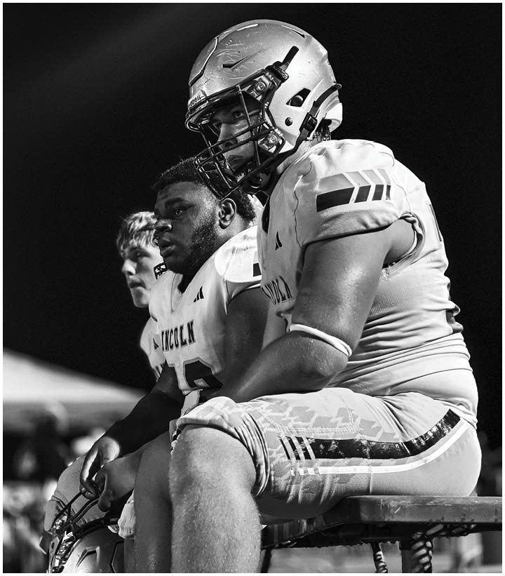 Three people in football uniforms and helmets are closely positioned, appearing focused and attentive during a game or practice. Description generated by AI