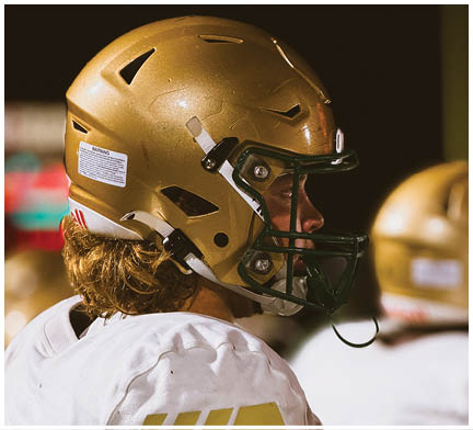 A person wearing a gold football helmet and white jersey is looking to the right during a nighttime outdoor event. Description generated by AI