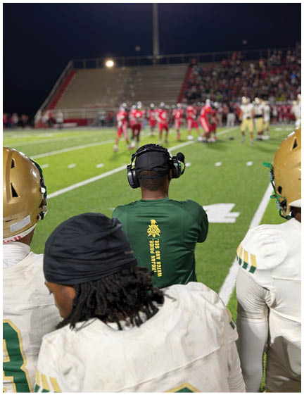 People wearing football helmets and uniforms stand on the sidelines while a coach in a headset watches players on the field during a nighttime game. Description generated by AI