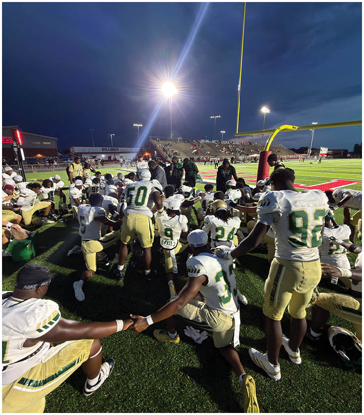 A group of people in football uniforms and coaches are gathered on a football field near a goalpost under bright stadium lights with spectators in the bleachers in the background. Description generated by AI