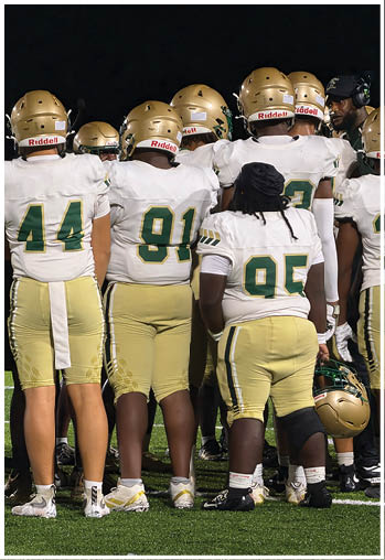 Four people wearing white football uniforms with gold helmets are gathered closely together on a dark field. Description generated by AI