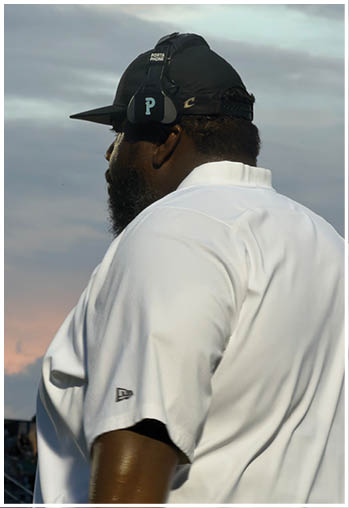A person wearing a gold football helmet and white jersey with green and gold accents is standing outdoors under stadium lights during dusk or evening. Description generated by AI