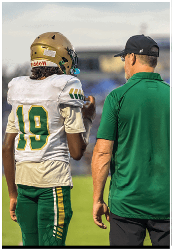 Person wearing a gold football helmet with a green faceguard and a white jersey against a dark blue background. Description generated by AI