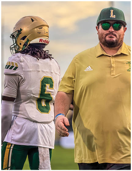 A person wearing a headset and cap is speaking closely to a person in a Lincoln sports jersey who has a serious expression. Description generated by AI