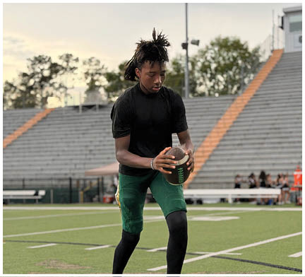 A person with dreadlocks wearing a black shirt and green pants is holding a football on a sports field with empty bleachers in the background. Description generated by AI