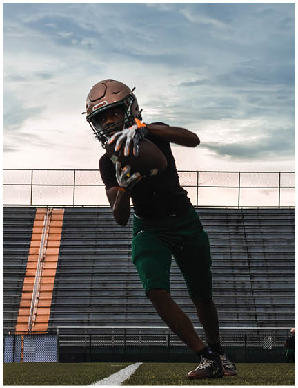 A person wearing a helmet and gloves is catching a football on a field with empty bleachers and a cloudy sky in the background. Description generated by AI