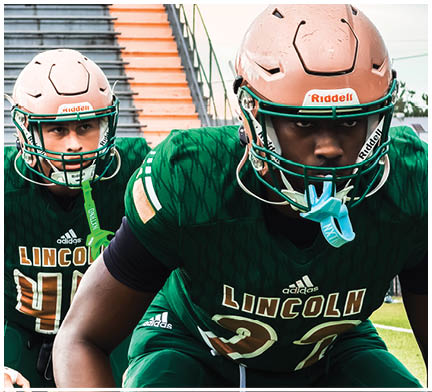 Two people wearing green football uniforms and helmets are positioned on a field with stadium stairs in the background. Description generated by AI