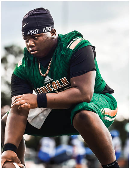 A person wearing a green sports uniform and black headband is crouched down holding a football on a field with blurred players in the background. Description generated by AI