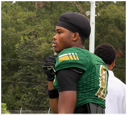 An adult wearing a green football uniform with the number 10 stands on the sidelines of a football field while another adult in a white shirt stands nearby. Description generated by AI