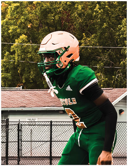 A person wearing a green football uniform and a peach helmet is standing on a field with trees and a building in the background. Description generated by AI