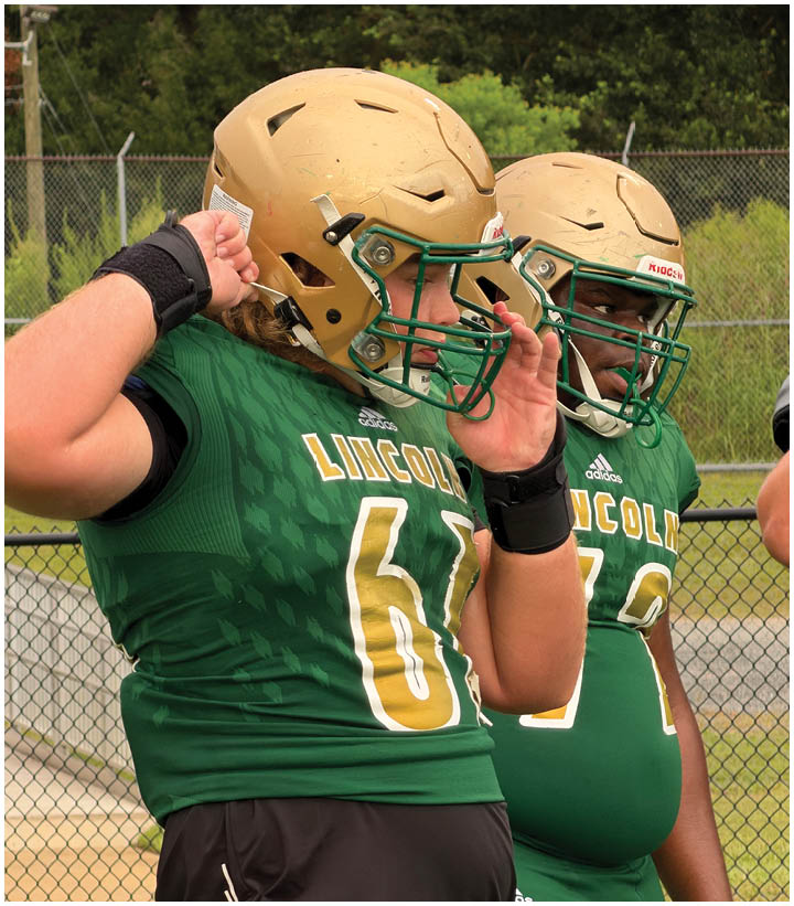 Two people wearing green football jerseys and gold helmets stand near a chain-link fence with trees in the background. Description generated by AI