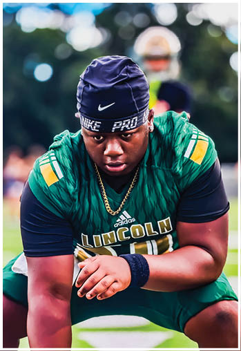 A person wearing a gold football helmet and white jersey with green and gold accents is standing outdoors under stadium lights during dusk or evening. Description generated by AI