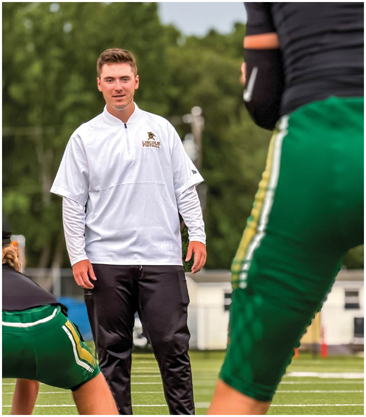A person wearing a white long-sleeve shirt and black pants stands on a football field while two people in green sports uniforms prepare for action nearby. Description generated by AI