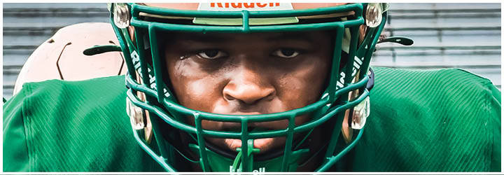 A person wearing a pink football helmet with a green faceguard and a green jersey is looking directly ahead on a sports field. Description generated by AI