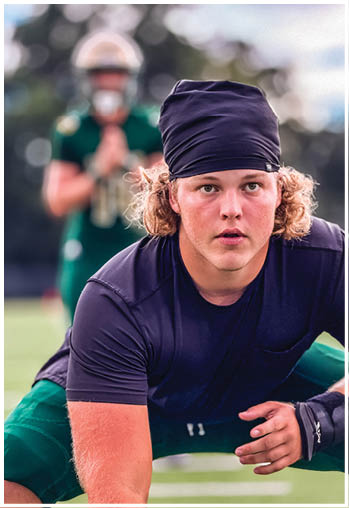 An adult wearing a dark head covering and shirt is crouched in a ready position on a sports field with another person in green sports gear blurred in the background. Description generated by AI