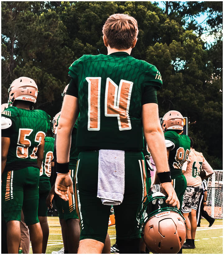People wearing green football uniforms stand on a field with one holding a helmet and a towel hanging from their waist. Description generated by AI
