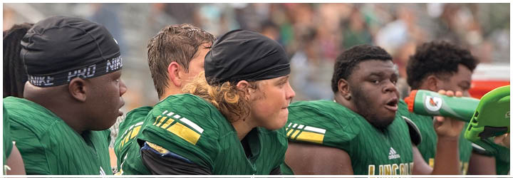 Several people wearing green sports jerseys and headbands are sitting on a bench with a blurred crowd in the background. Description generated by AI