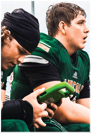 Two people in green football uniforms are sitting closely, with one holding a green device and the other looking forward. Description generated by AI