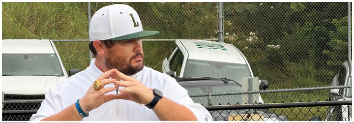 A white and green baseball cap with the letter L on it is placed in front of a chain-link fence with dense green foliage behind it. Description generated by AI