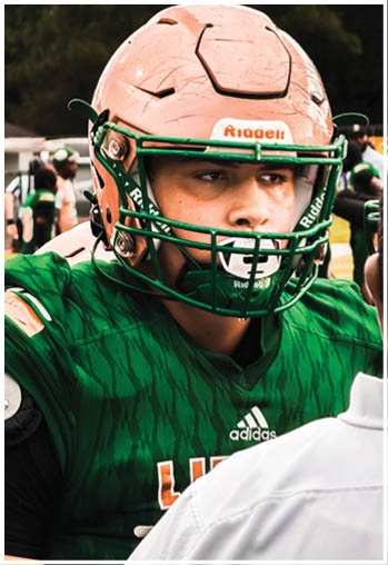 A person wearing a green Adidas football jersey and a pink Riddell helmet is focused on the game with other players blurred in the background. Description generated by AI