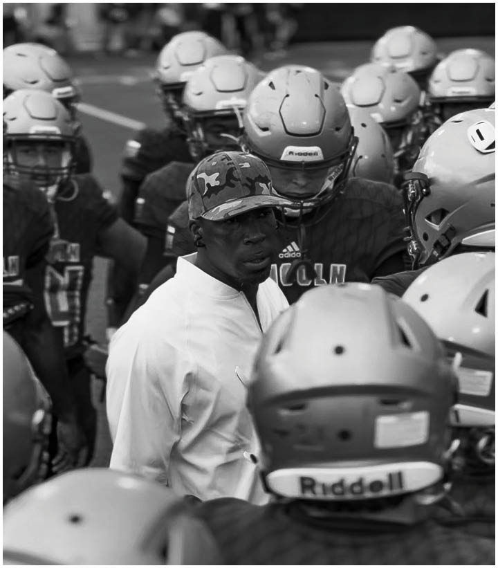 A person wearing a camouflage cap and white shirt is surrounded by a group of people in football helmets and uniforms on a field. Description generated by AI