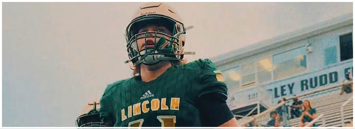 A person wearing a green sports jersey and a football helmet is standing outdoors near a building with windows. Description generated by AI