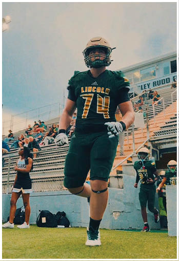 An adult wearing a football uniform with the number 74 and the word Lincoln is walking on a field near bleachers with other people in sports attire. Description generated by AI