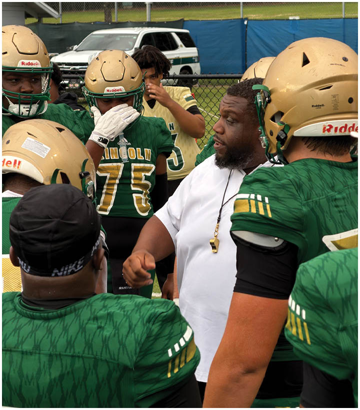 A group of people in green football uniforms and gold helmets are gathered around a person in a white shirt who appears to be giving instructions on a sports field. Description generated by AI
