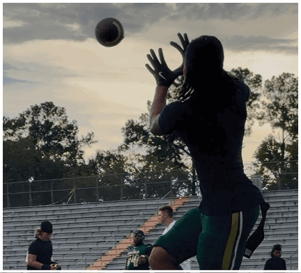 People wearing athletic clothing are practicing catching a football on a field with empty bleachers and trees in the background. Description generated by AI