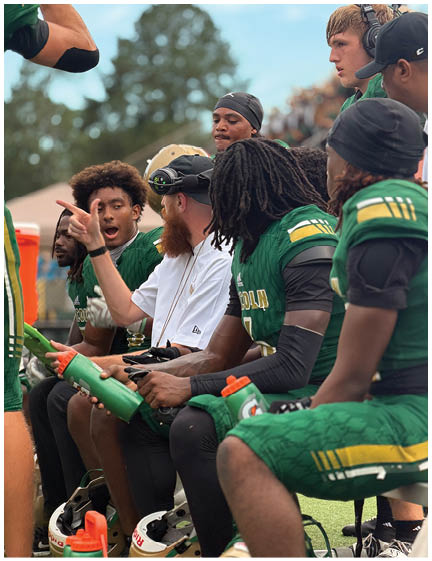 People in green sports uniforms sit on a bench during a game while one person in a white shirt and cap gestures and talks to them. Description generated by AI