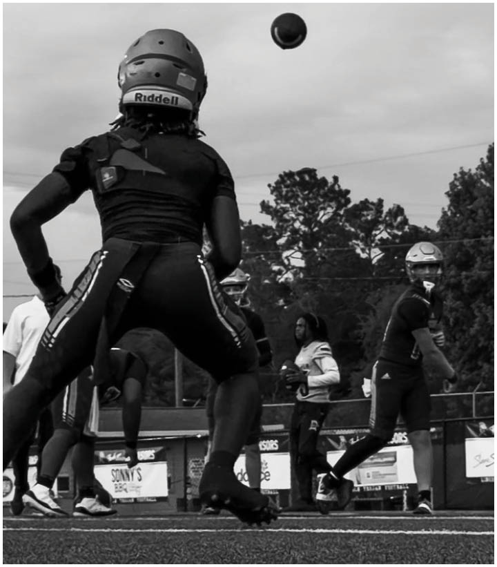People wearing helmets and sports gear are playing football on a field with trees and a fence in the background. Description generated by AI