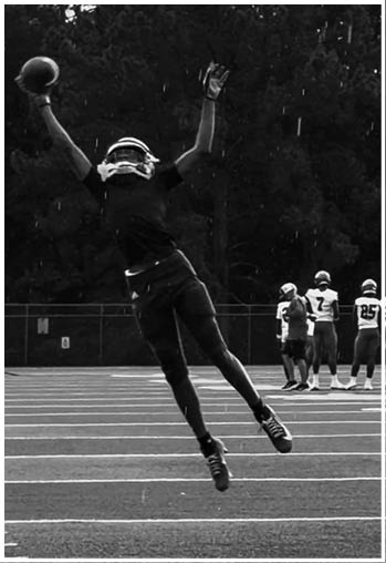 A person in sports gear is leaping to catch a football on a field while several other people in uniforms stand in the background. Description generated by AI