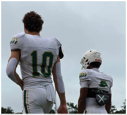 Two people wearing white football uniforms with green and yellow accents are standing outdoors against a cloudy sky, one with a helmet and the other without. Description generated by AI